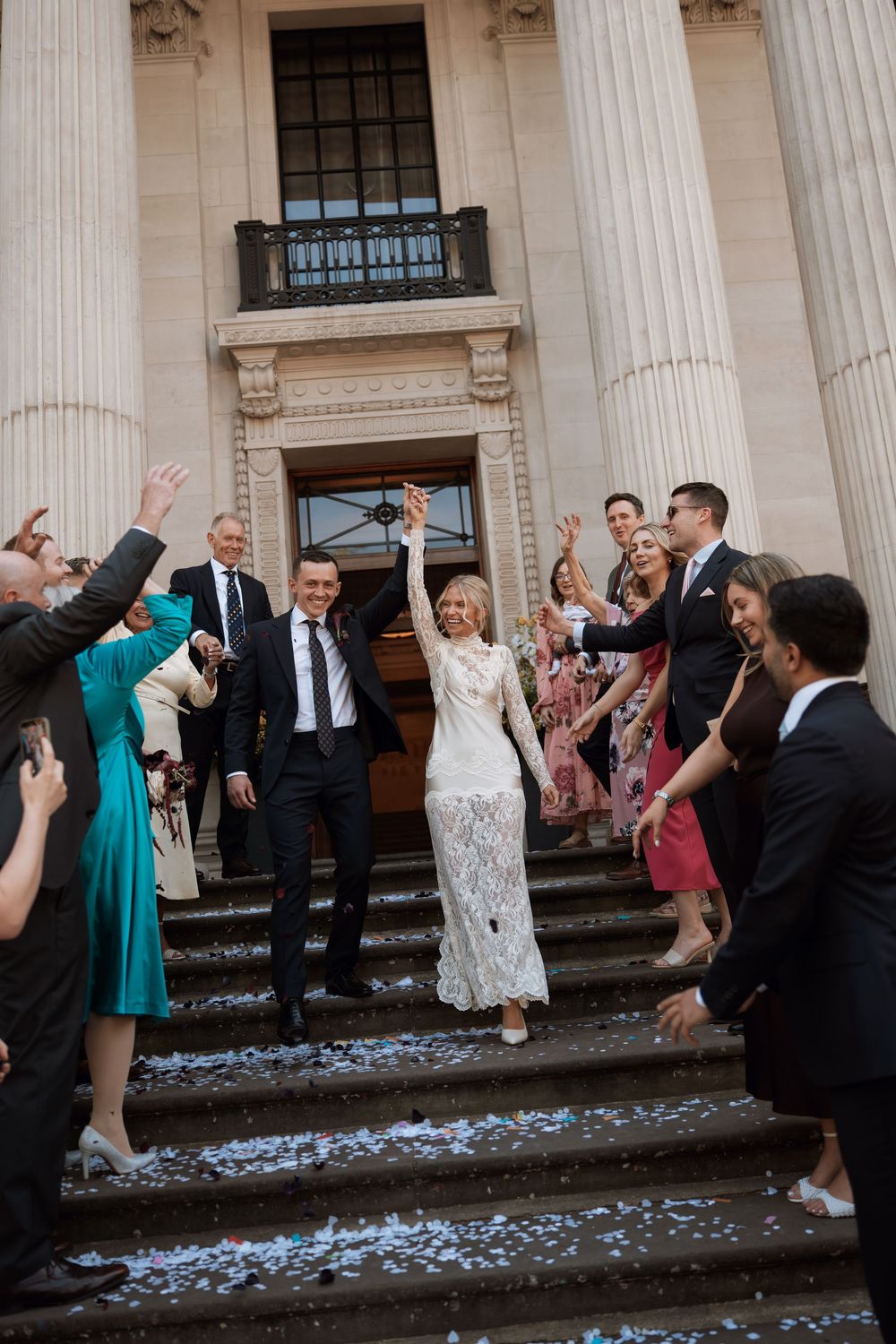 Bride and groom celebrate on steps of grand building, surrounded by guests throwing white confetti at joyful wedding ceremony.