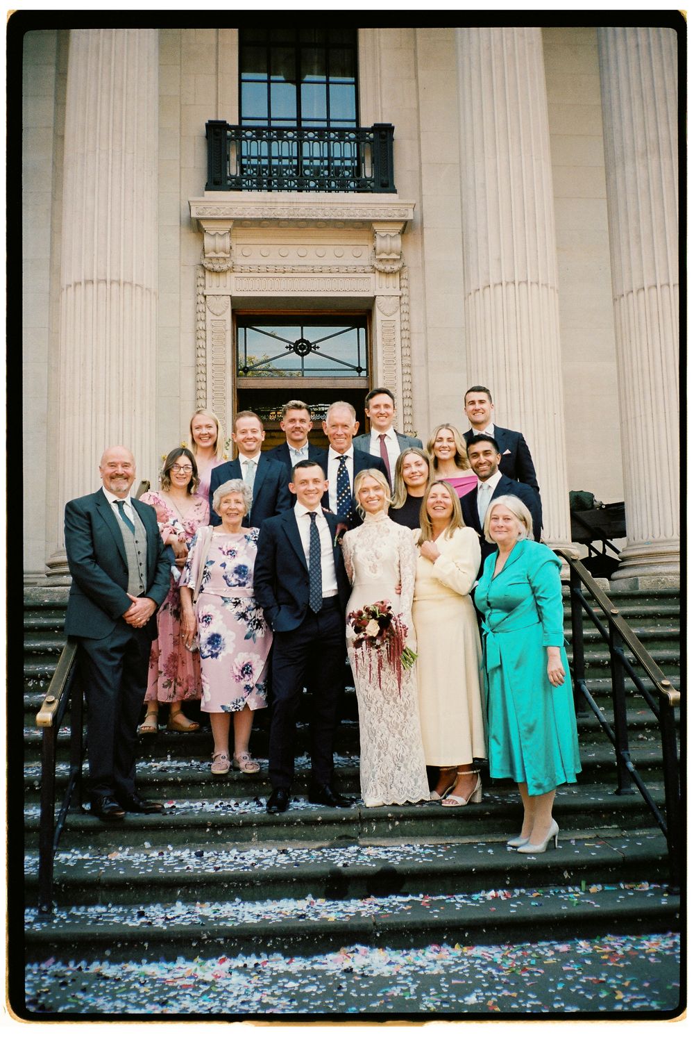 Wedding party posing on steps of grand building, bride in lace dress holding bouquet, guests in formal attire.