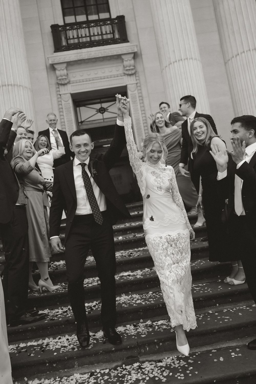 Black and white photo of a joyful newlywed couple raising hands on church steps, surrounded by cheering guests throwing confetti.