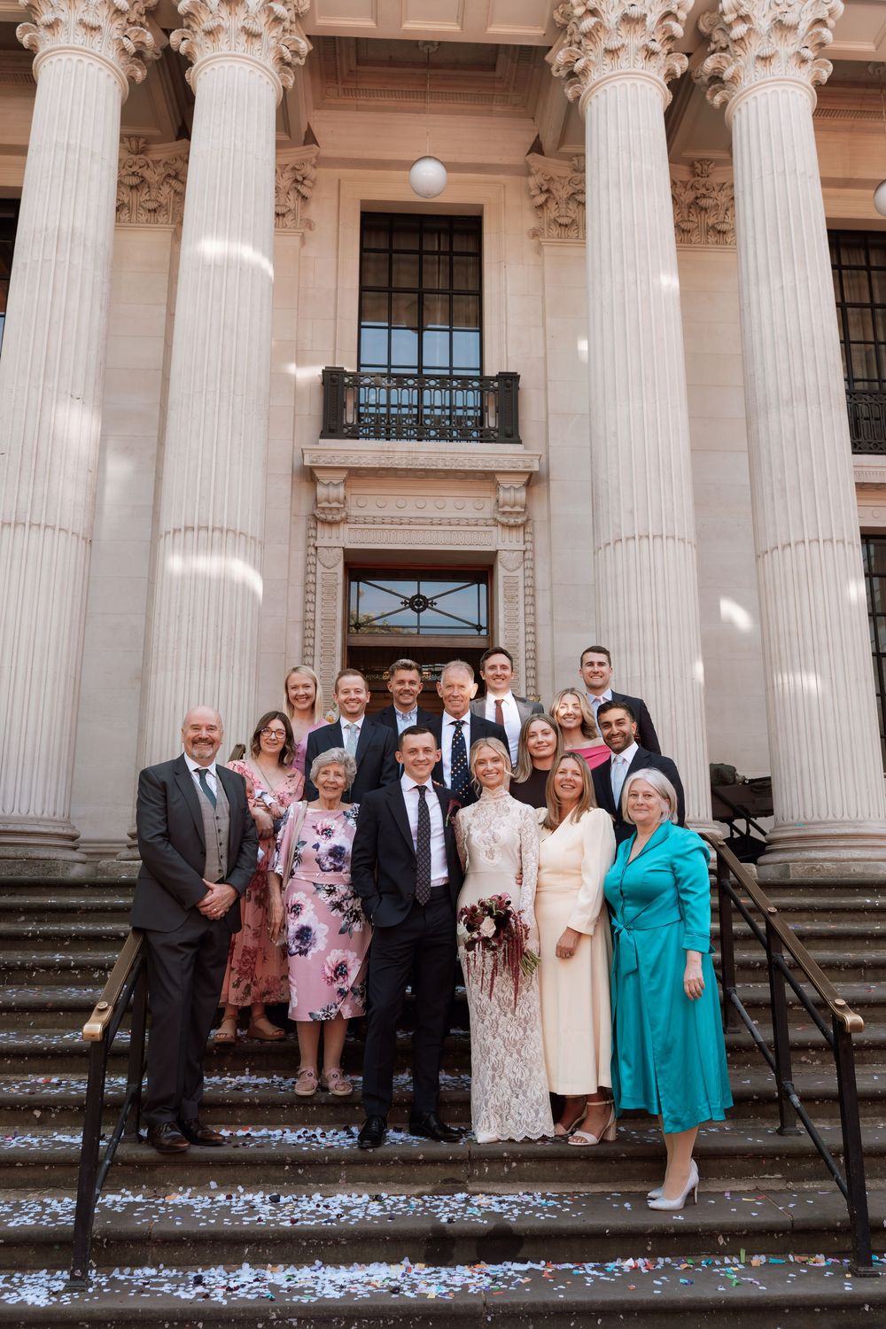 Wedding party posing on steps of grand neoclassical building with columns, bride in white dress, guests in formal attire.