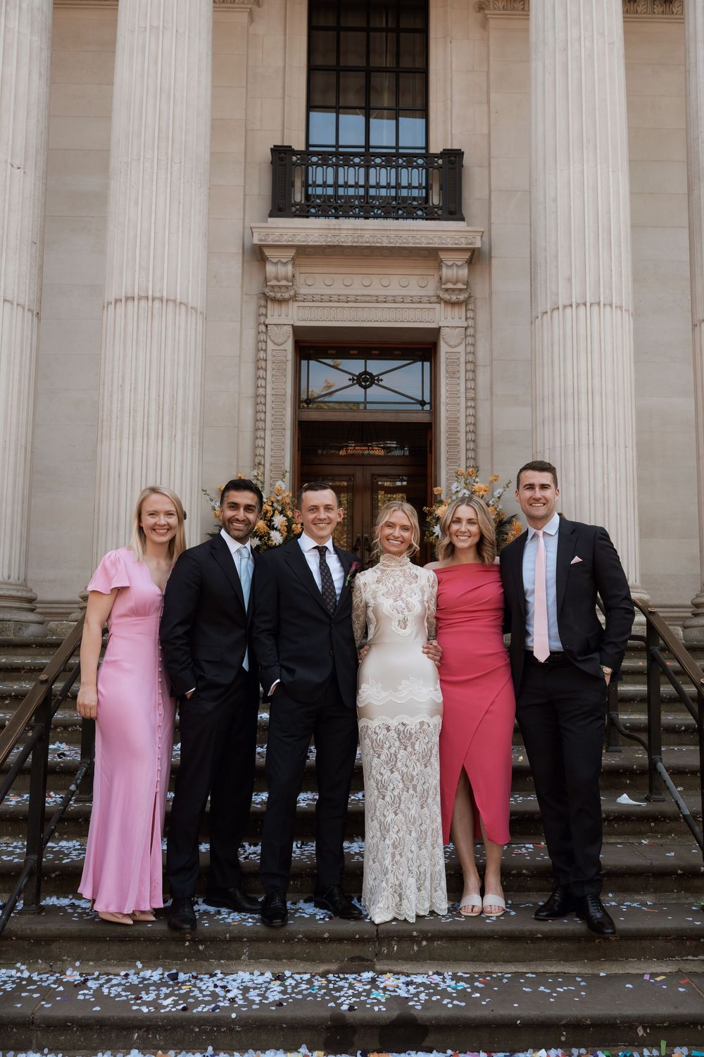 Group of six elegantly dressed people posing on steps outside a grand building at a wedding celebration.