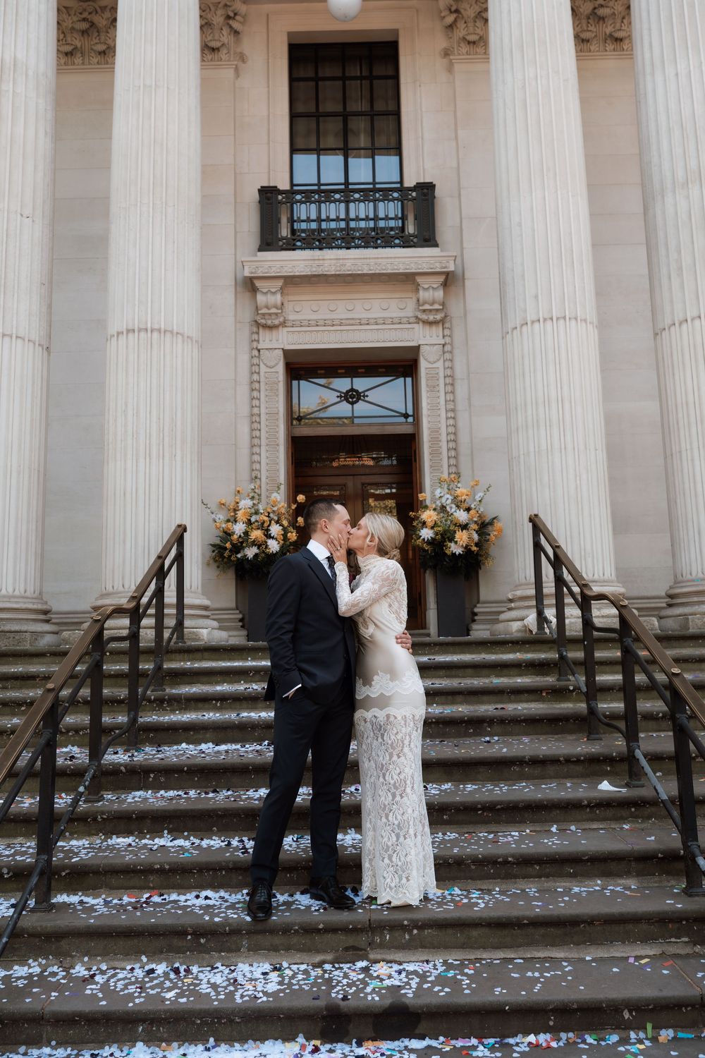 Newlywed couple kissing on steps of grand neoclassical building with floral arrangements and confetti.