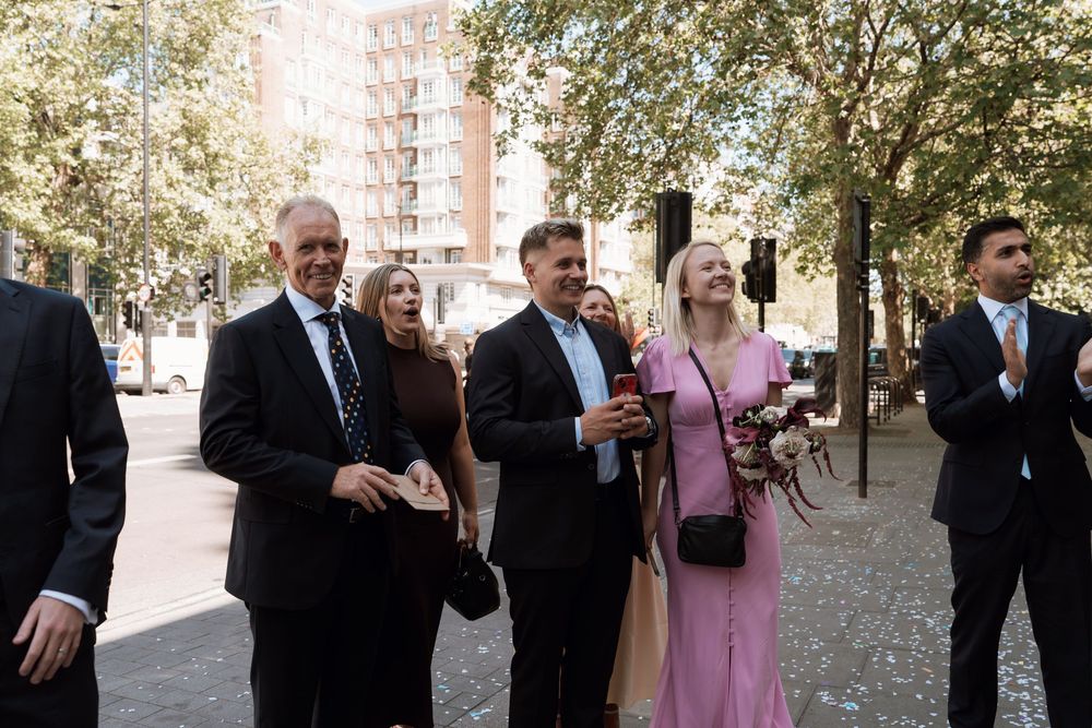 Group of elegantly dressed people laughing and celebrating outdoors at a wedding reception on a sunny day.