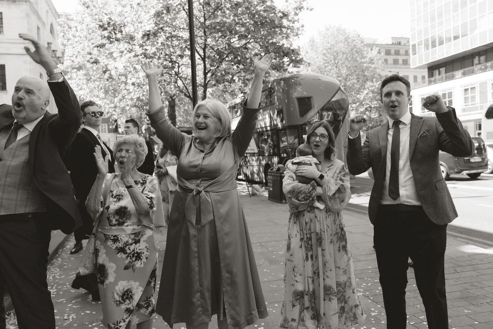 Black and white photo of joyful wedding guests celebrating outdoors, raising arms and cheering on a city street.