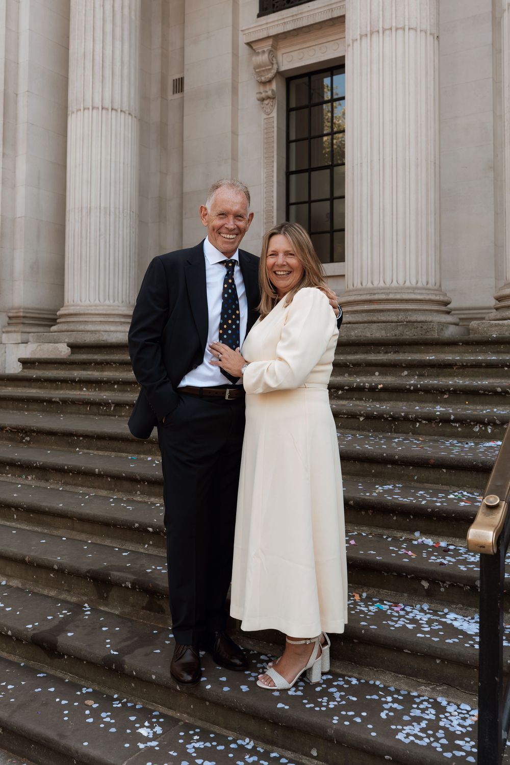 Couple smiling on steps of a grand building with columns, woman in white dress, man in dark suit, confetti scattered around.