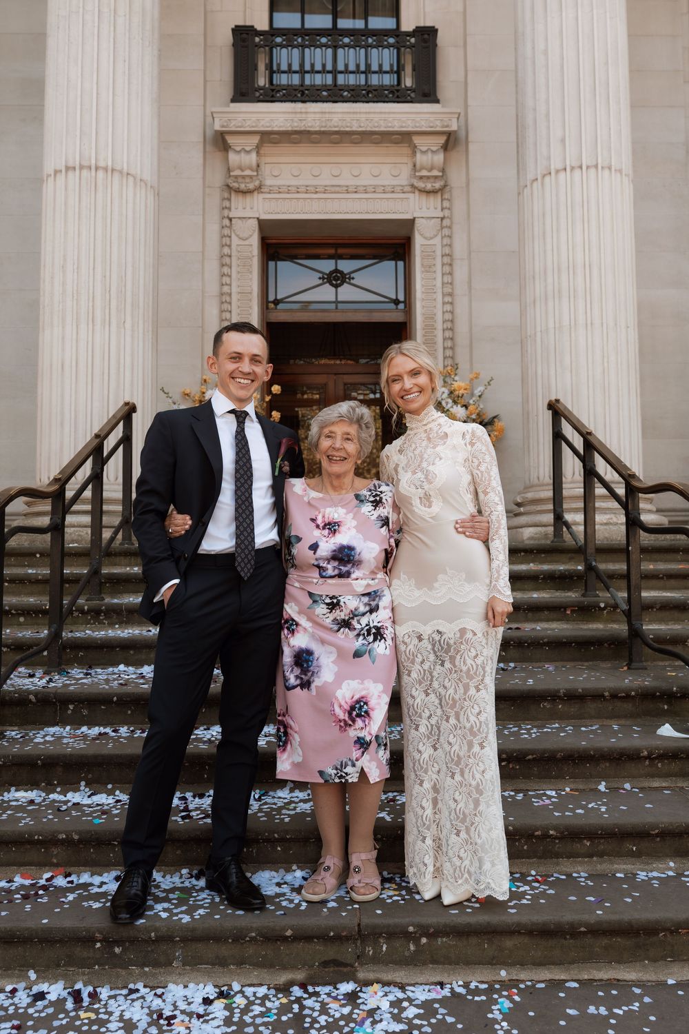 Bride and groom posing with elderly woman on steps of elegant building with confetti.