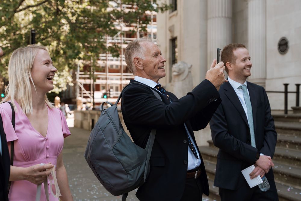 Three professionals smiling outdoors, one man taking a photo with smartphone near a historic building.