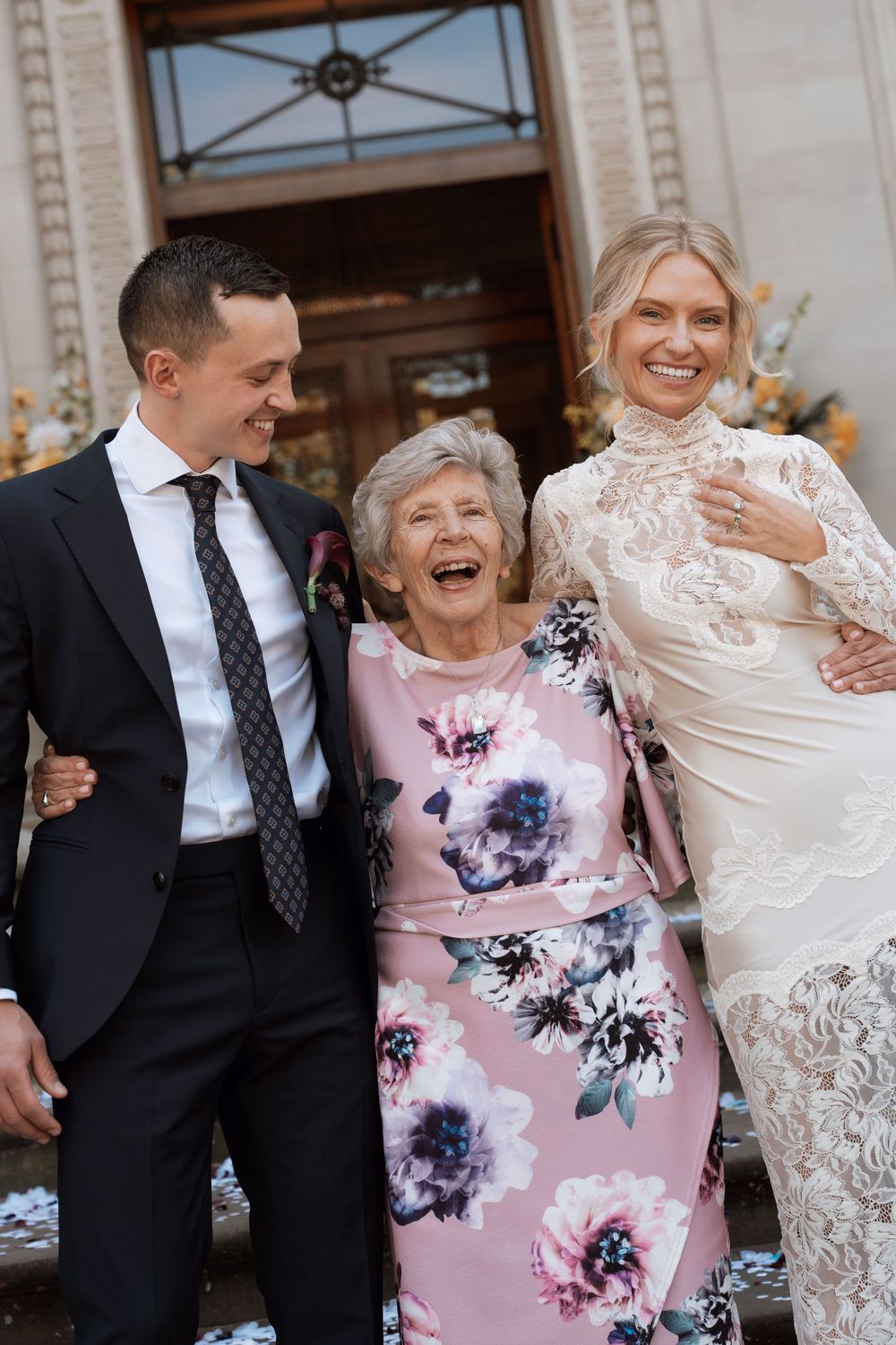 Bride in lace gown and groom in black suit laugh with elderly woman in floral dress outside wedding venue.