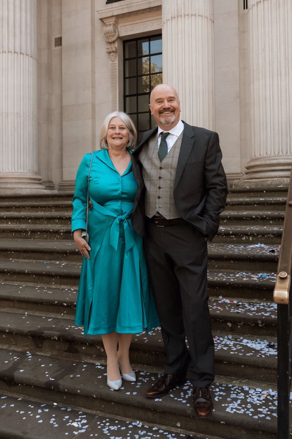 Smiling couple in formal attire stand on stone steps with confetti, woman in teal dress, man in grey vest and suit.