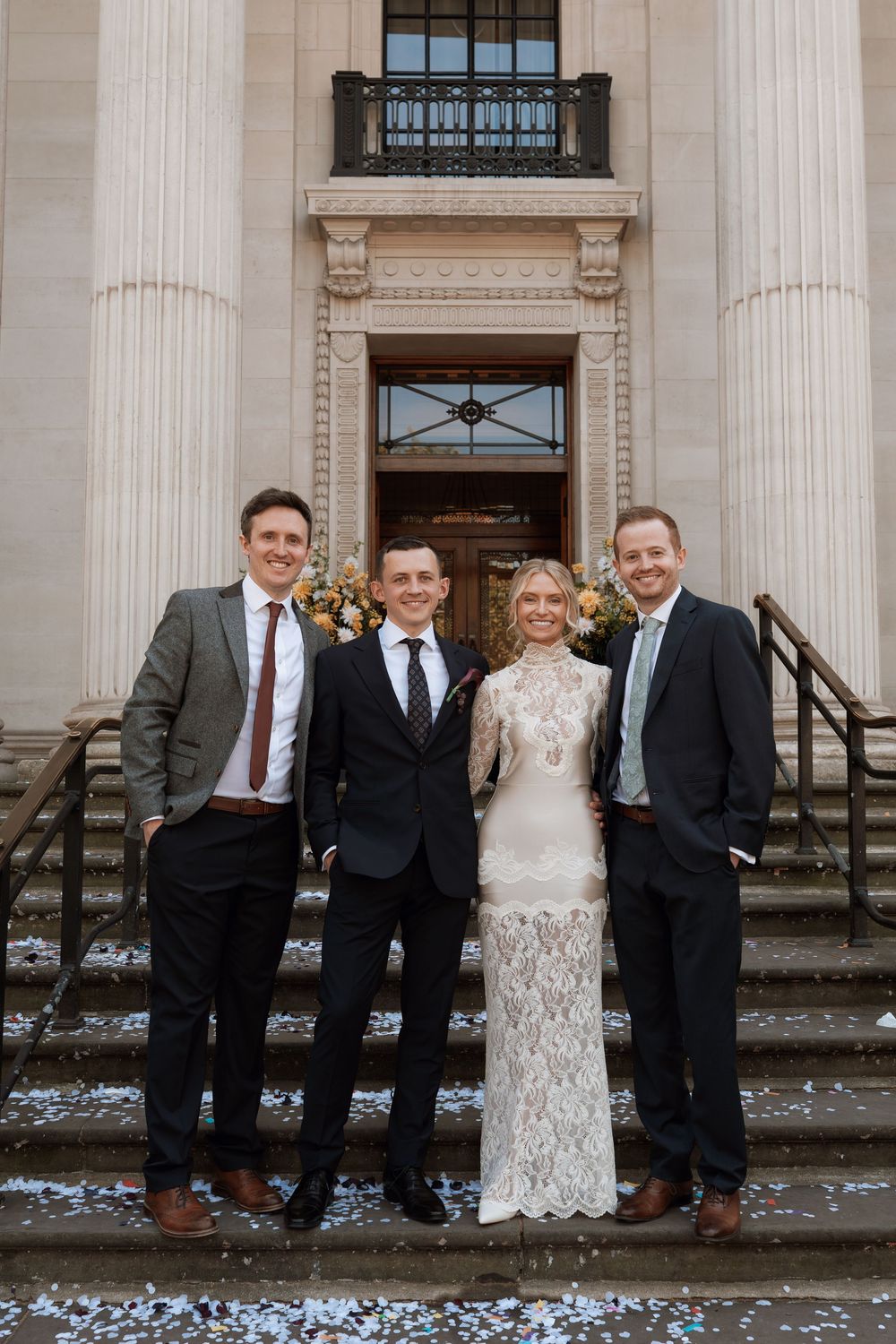Bride in lace gown poses with three suited groomsmen on steps of elegant neoclassical building with confetti.