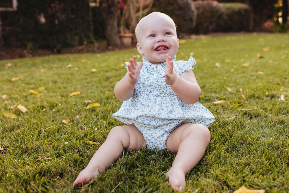 Happy baby girl in a floral romper clapping hands while sitting on green grass outdoors.