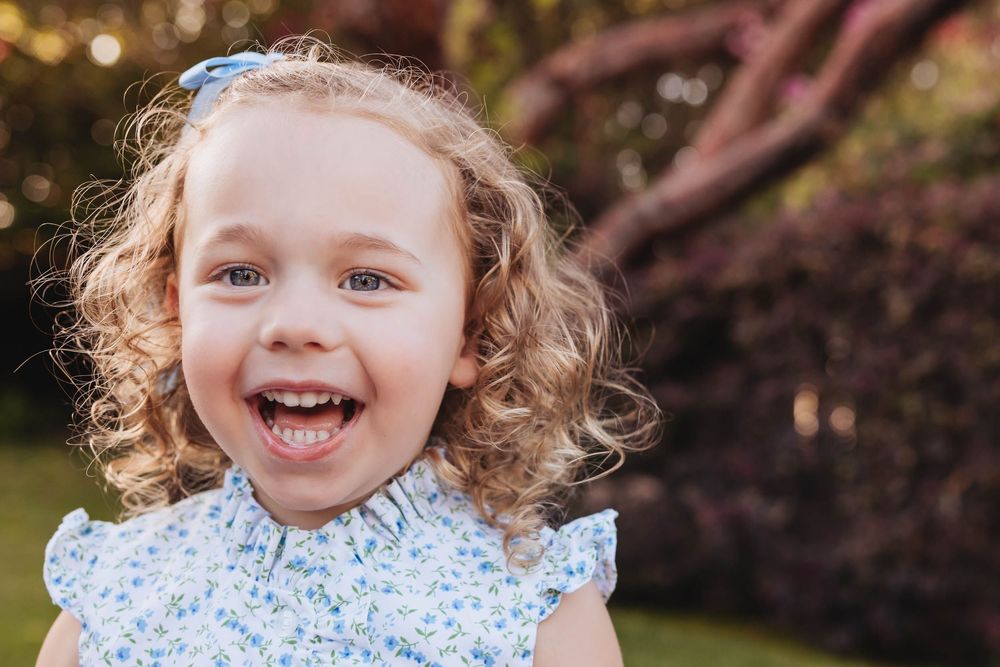 A joyful young girl with curly blonde hair and blue eyes smiles brightly outdoors in a floral dress.