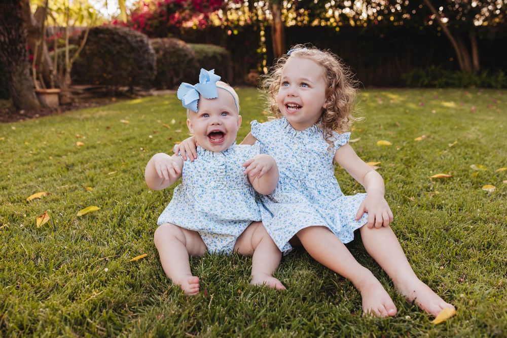 Two joyful baby girls in matching blue floral outfits sitting on green grass, laughing outdoors.