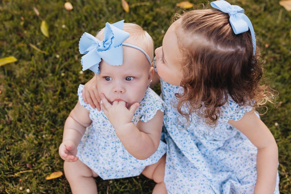 Two young sisters in matching blue floral dresses sit on grass, older girl kissing baby sister wearing a blue bow.