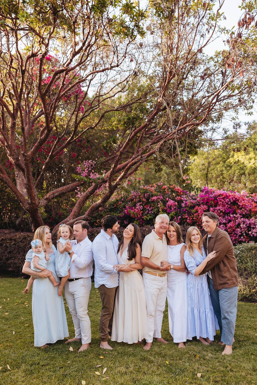 Large family group posing outdoors on green lawn surrounded by blooming pink azaleas and trees in a garden setting.
