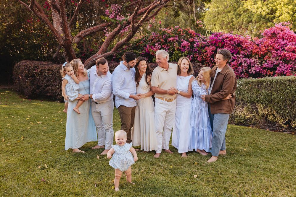 Large family group smiling outdoors on green lawn with colorful flowering bushes, toddler walking in foreground while on vacation