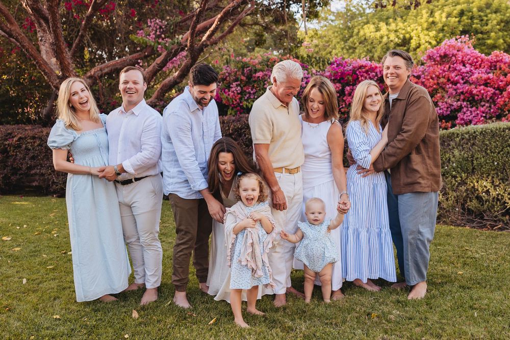 Large family group portrait outdoors on green lawn with blooming pink flowers, multiple generations smiling together.