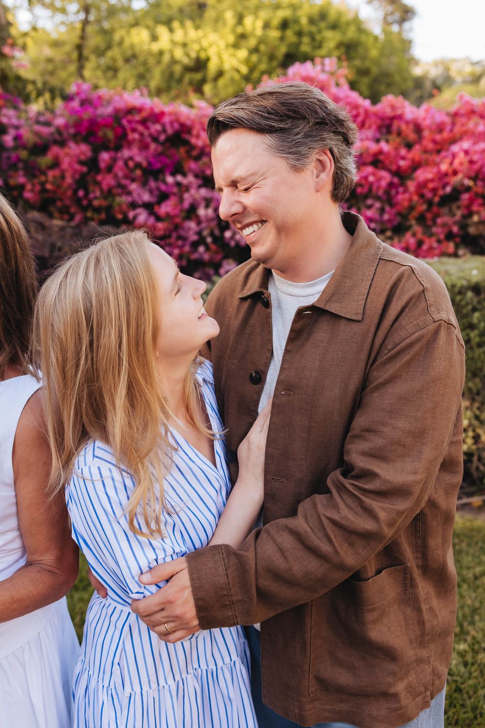 A smiling couple embraces outdoors with vibrant pink flowering bushes in the background on a sunny day.