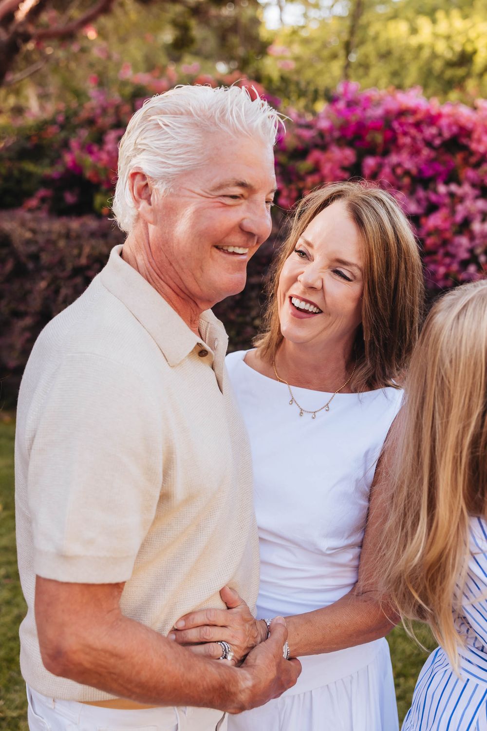 A smiling couple laughs together outdoors with vibrant pink flowering bushes in the background.
