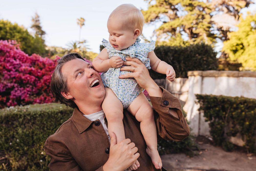 A laughing man holds a curious baby in a floral outfit outdoors near colorful bushes.