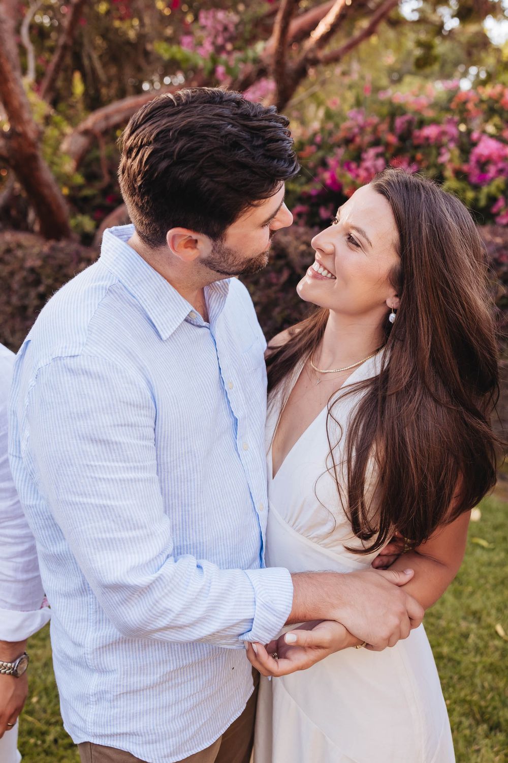 A couple shares a romantic moment outdoors, smiling at each other near blooming pink flowers.
