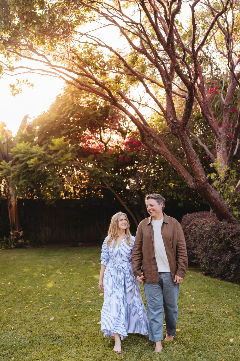 Couple holding hands walking barefoot on green grass at golden hour in a lush garden setting.