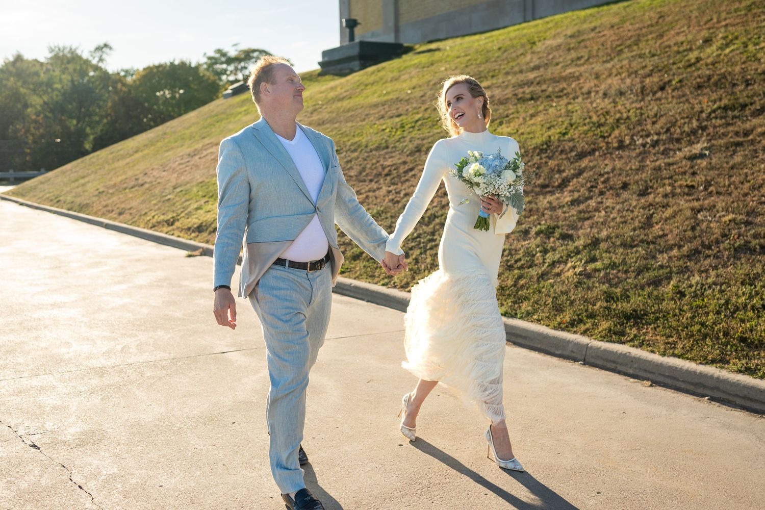 Bride and groom holding hands walking outdoors on sunny day, woman in white dress with bouquet, man in light blue suit.