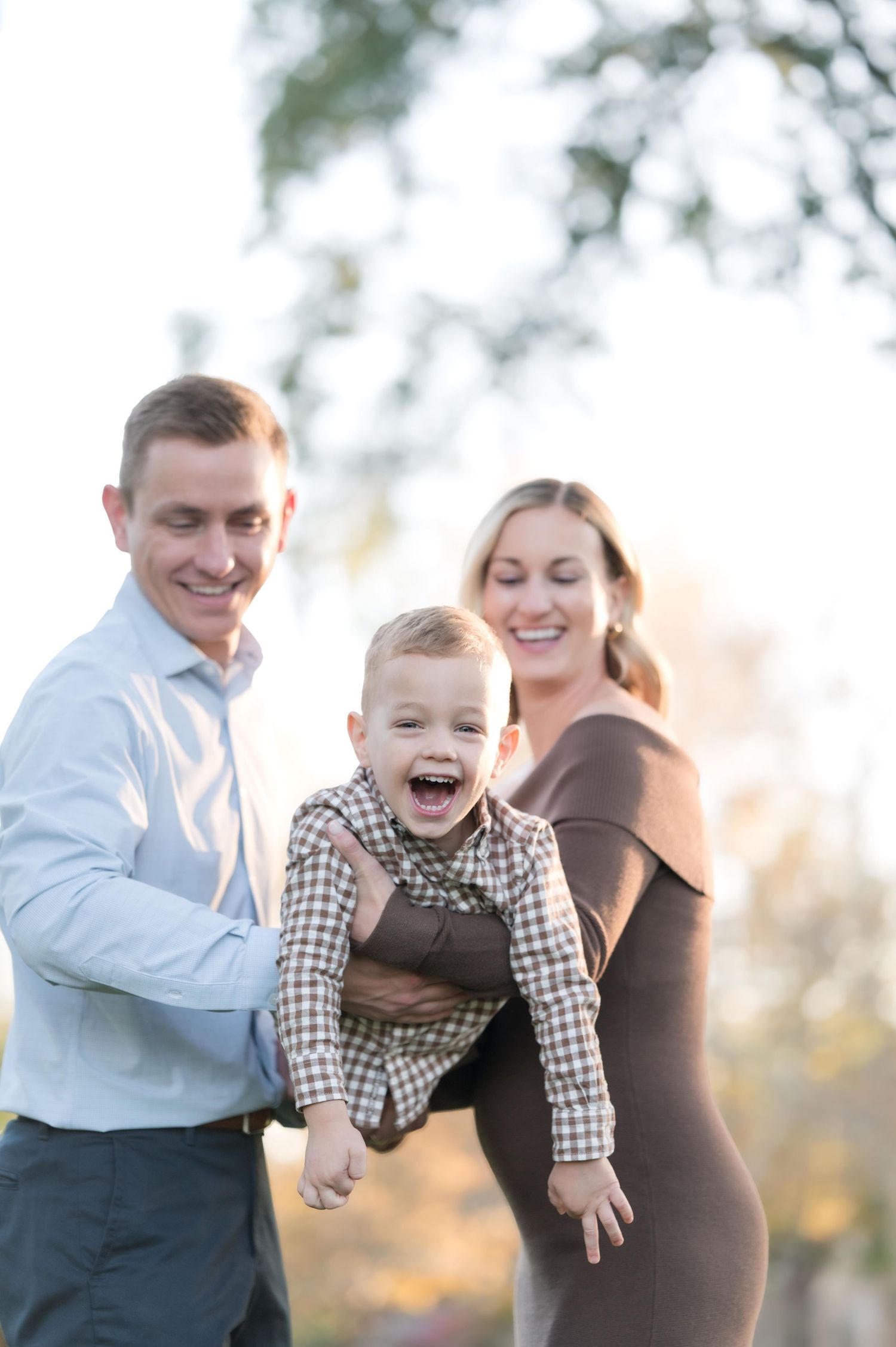 A joyful family photo with parents smiling as they lift their laughing toddler outdoors in warm autumn light.