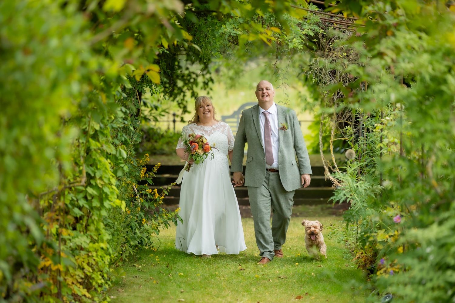 Bride and groom walk through a lush green garden arch with a golden dog on their wedding day. Casual Captures UK