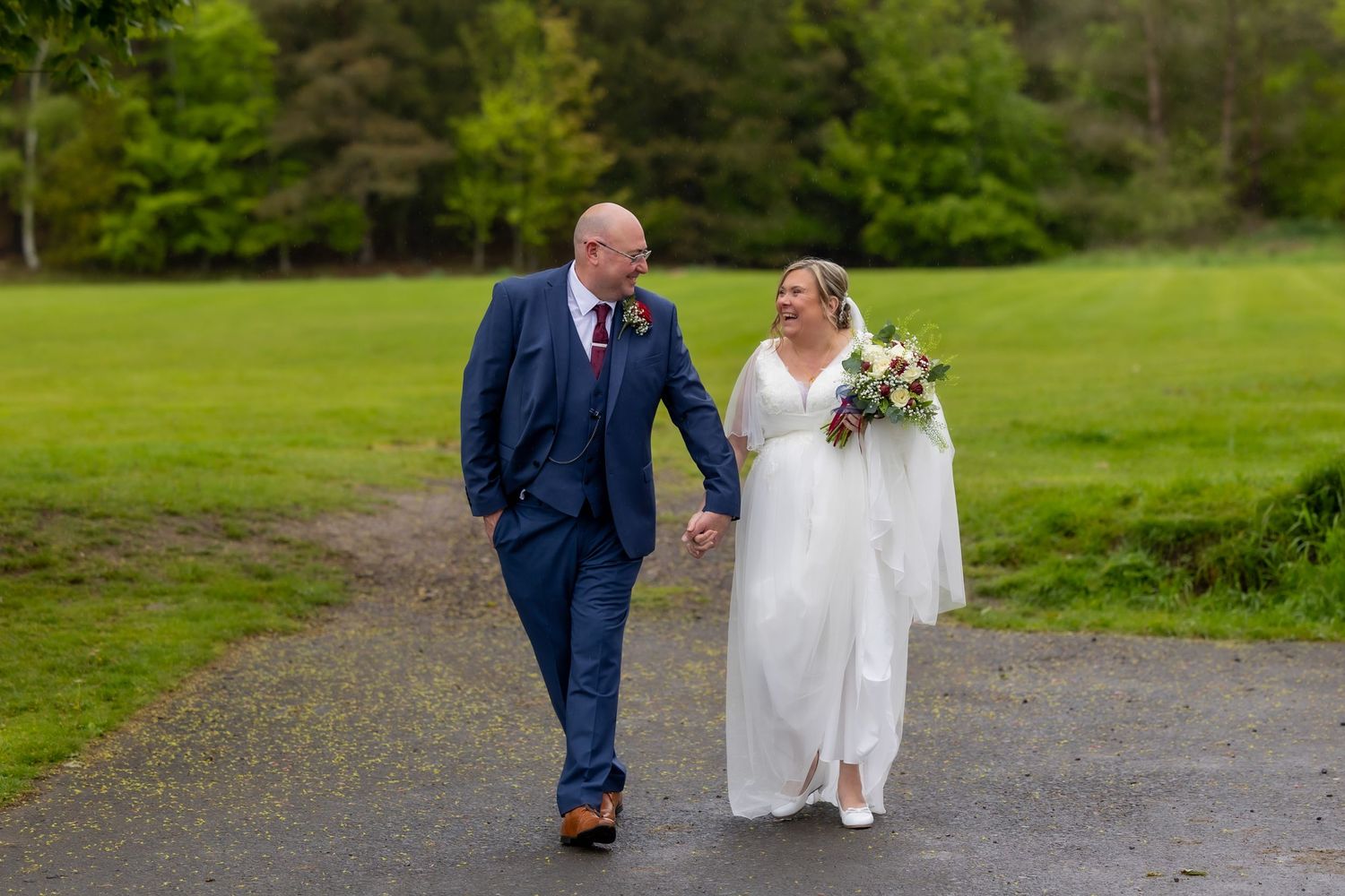 Bride and groom walk hand-in-hand on a path surrounded by lush green grass on their wedding day. Casual Captures UK