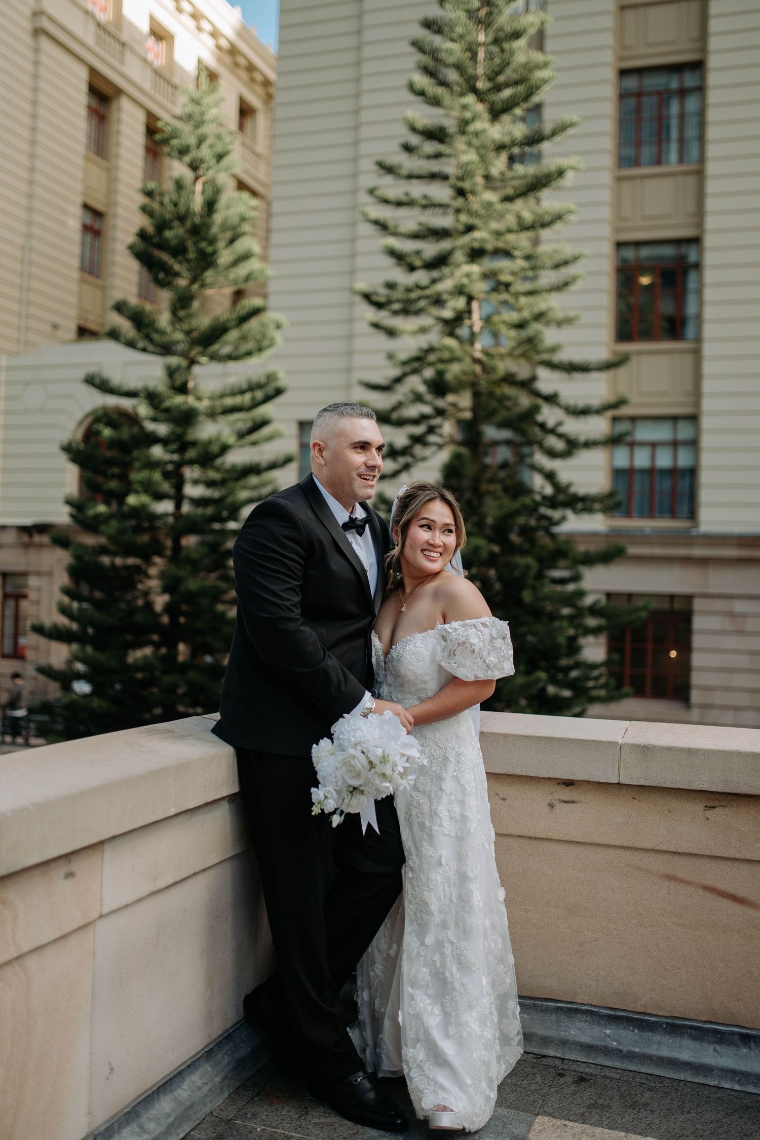 Bride in off-shoulder lace gown holds white bouquet beside groom in black tuxedo on rooftop terrace.