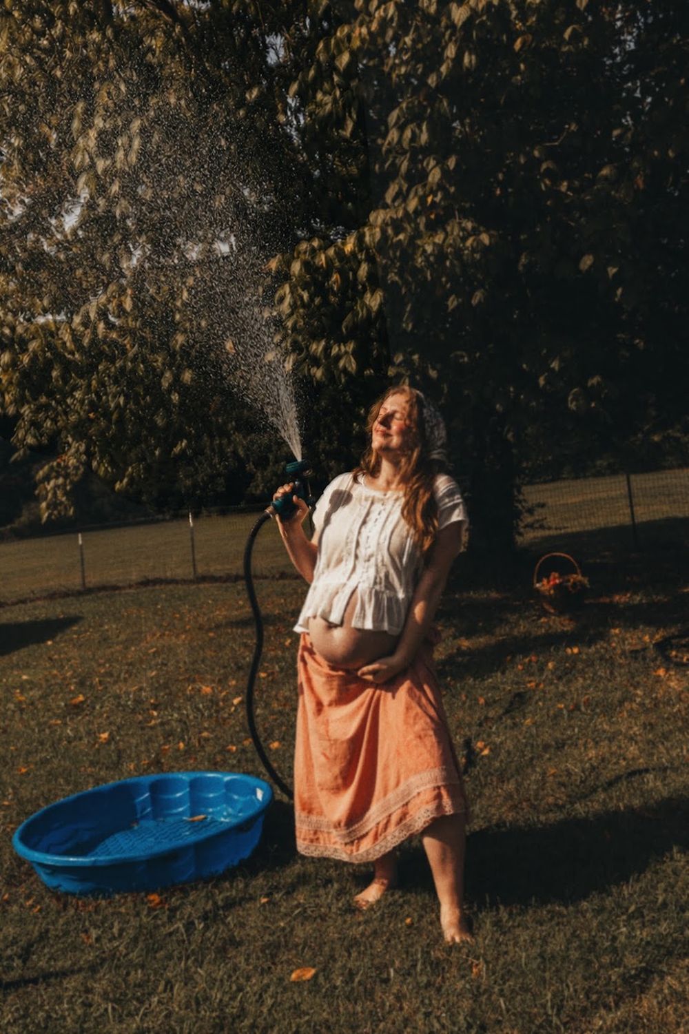 Pregnant woman in white top and orange skirt spraying garden hose outdoors near blue kiddie pool on sunny day in Knoxville, TN.