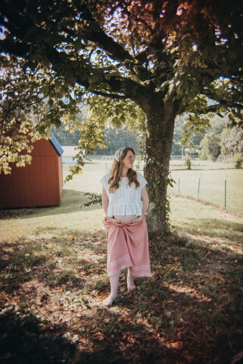 Pregnant woman in white top and pink skirt standing under a tree in a sunny green field near a red barn in Knoxville, TN.