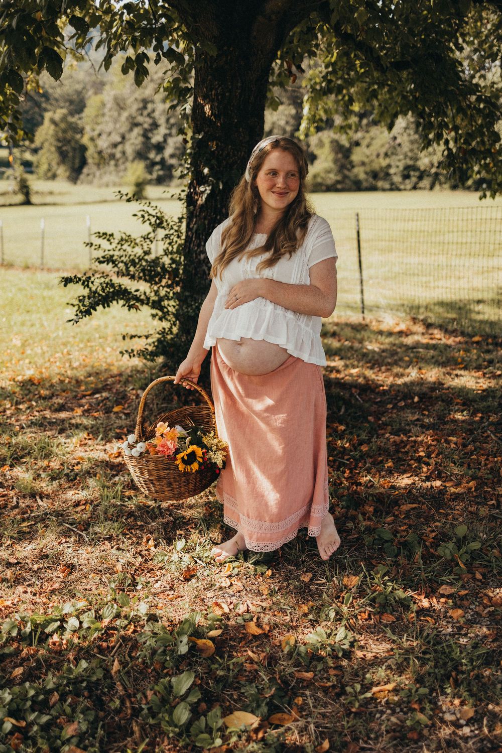 Pregnant woman in white top and pink skirt holding flower basket under tree in sunny park in Knoxville, TN.