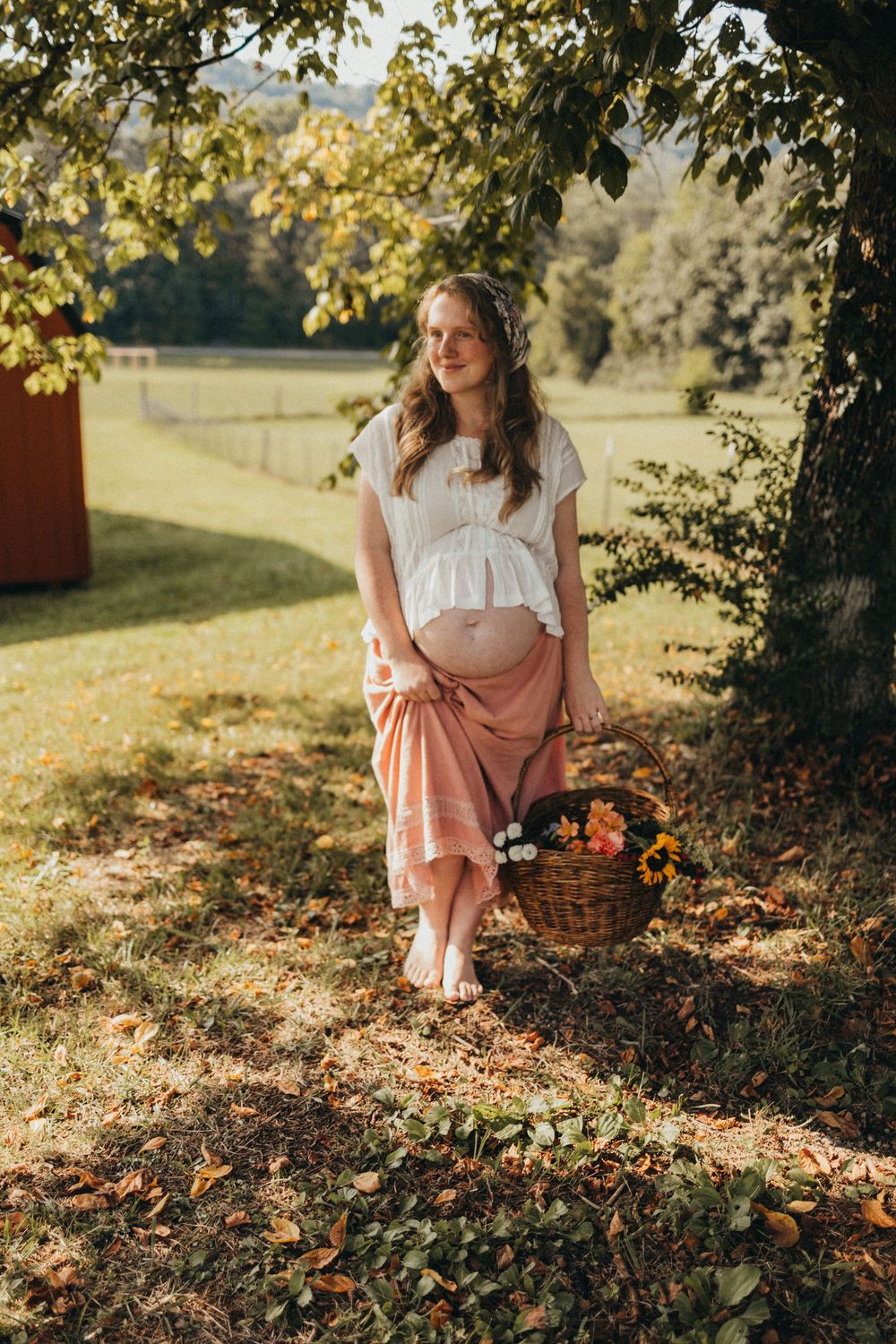 Pregnant woman in white top and pink skirt holding baby bump under tree with flower basket in sunny garden in Knoxville, TN.