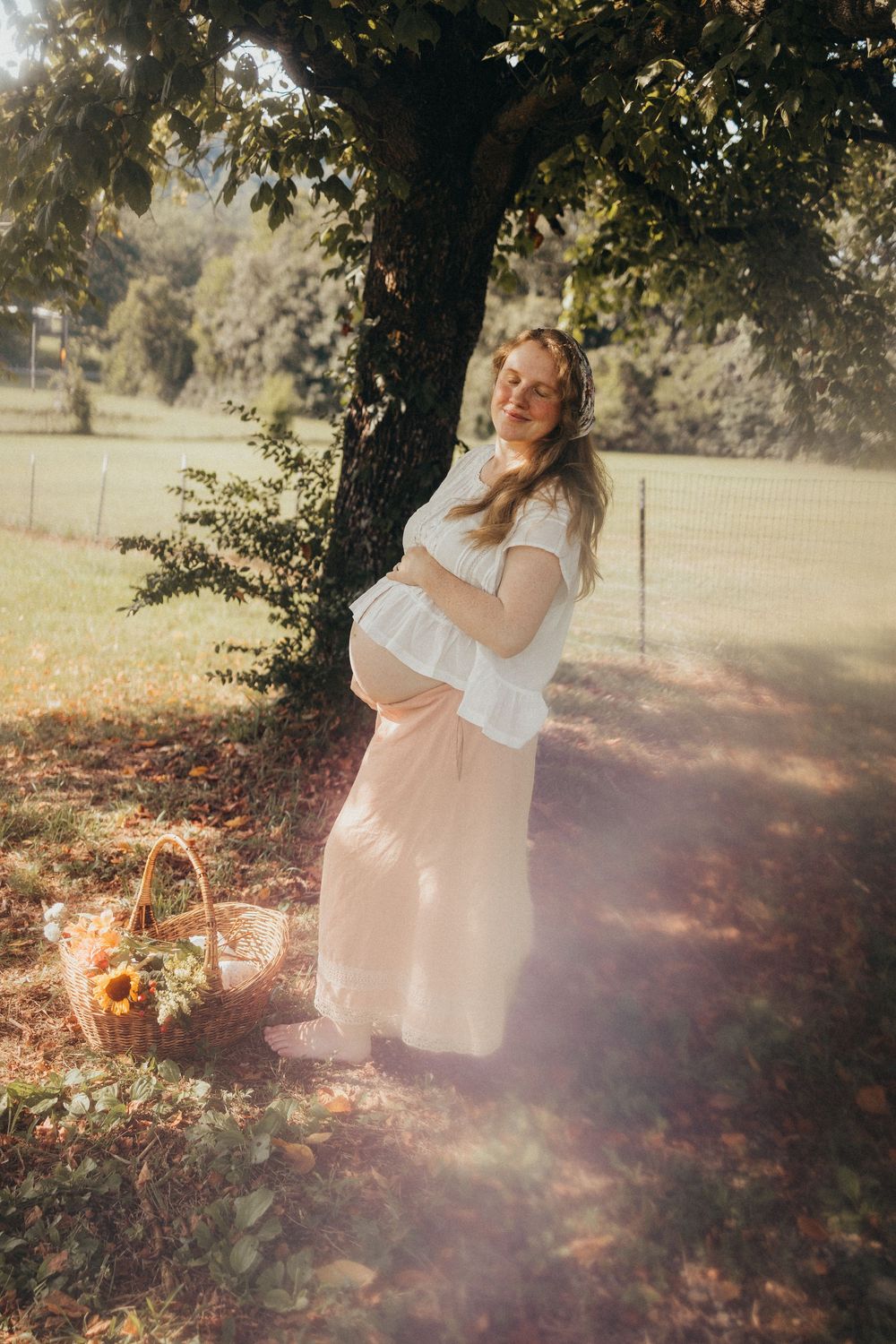 Pregnant woman in white top and pink skirt cradling her belly under a tree in a sunlit park with a flower basket in Knoxville, TN.