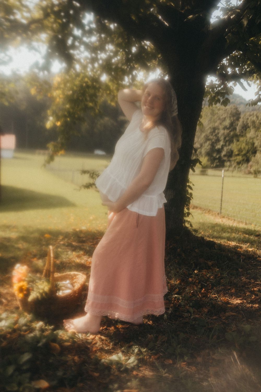Pregnant woman in white top and pink maxi skirt standing under a tree in a sunlit garden in Knoxville, TN.