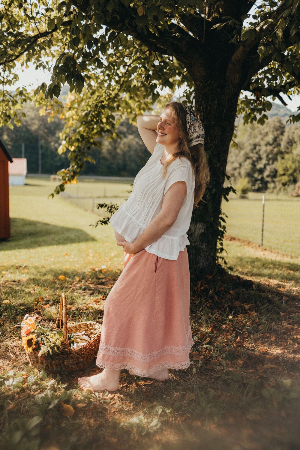 Smiling pregnant woman leaning against tree in white blouse and pink maxi skirt with wicker basket outdoors in Knoxville, TN.