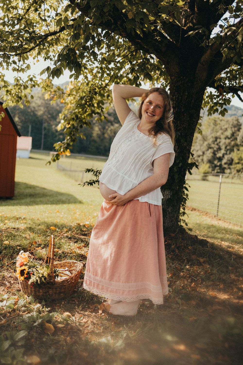 Smiling pregnant woman in white top and pink skirt leaning against a tree with a flower basket nearby in Knoxville, TN.