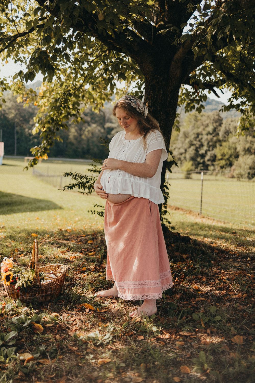 Pregnant woman in white blouse and pink skirt standing under a tree in a sunny garden, cradling her baby bump in Knoxville, TN.