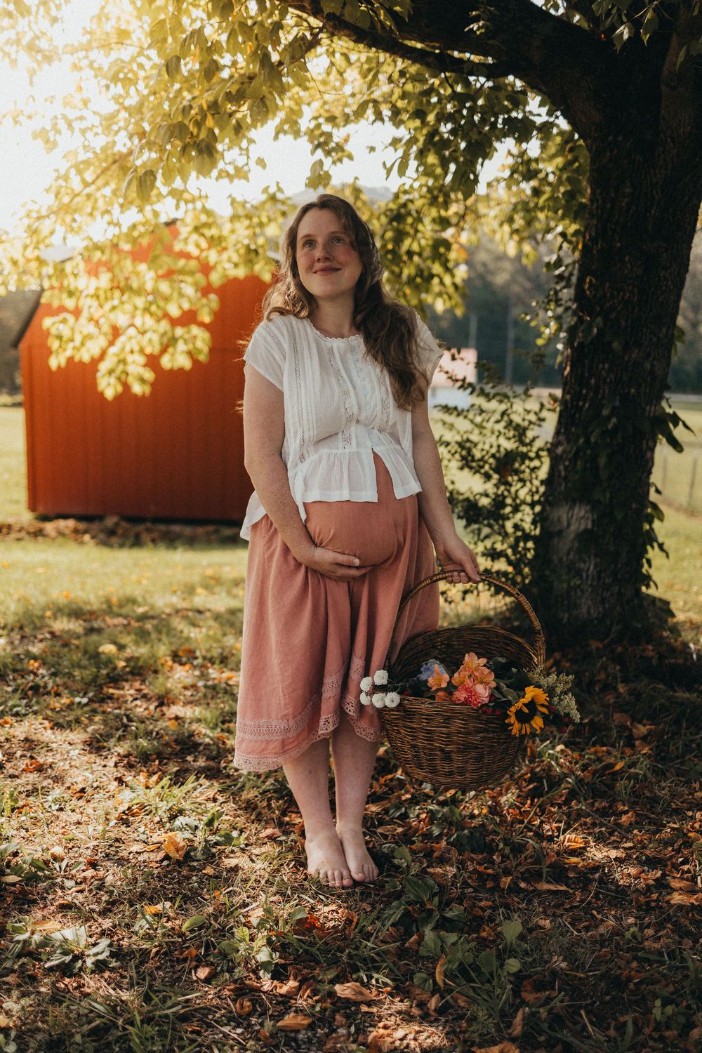Pregnant woman in white top and pink skirt holding baby bump beside a tree with a flower basket at golden hour in Knoxville, TN.