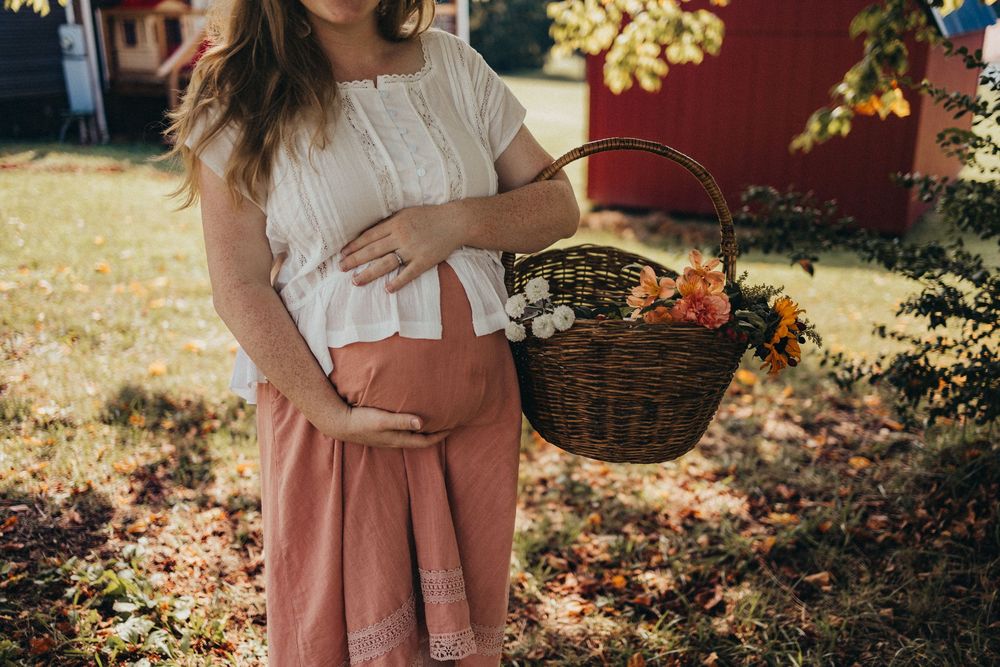 Pregnant woman in white top and pink skirt holding a wicker basket with fall flowers outdoors in sunlight in Knoxville, TN.