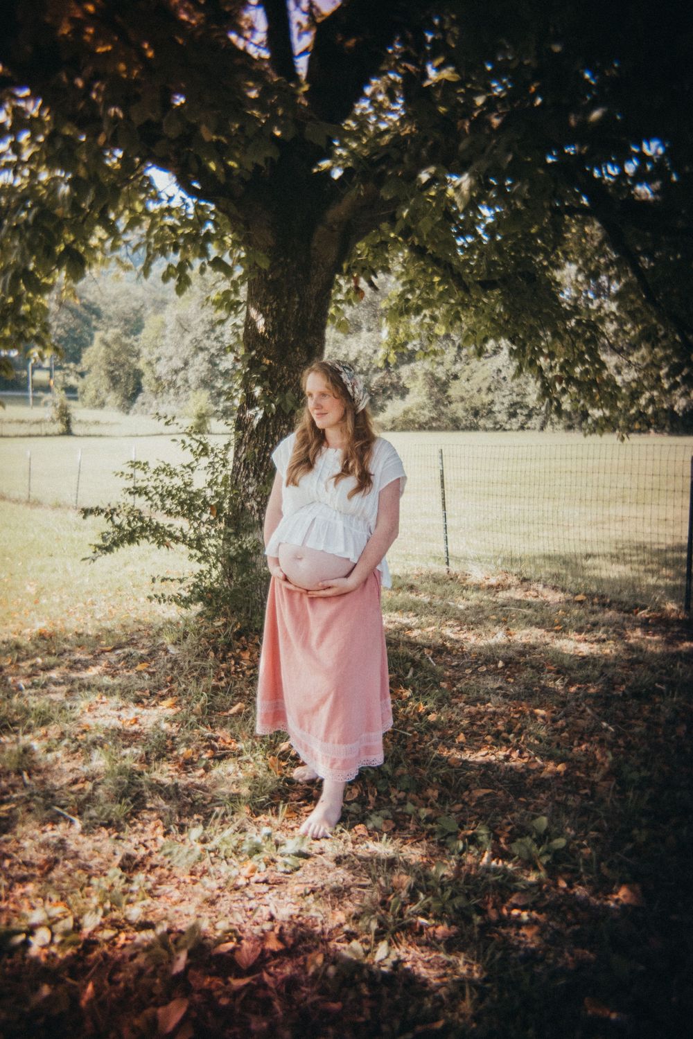 Pregnant woman in white top and pink skirt standing under a tree in a sunny park, cradling her baby bump in Knoxville, TN.