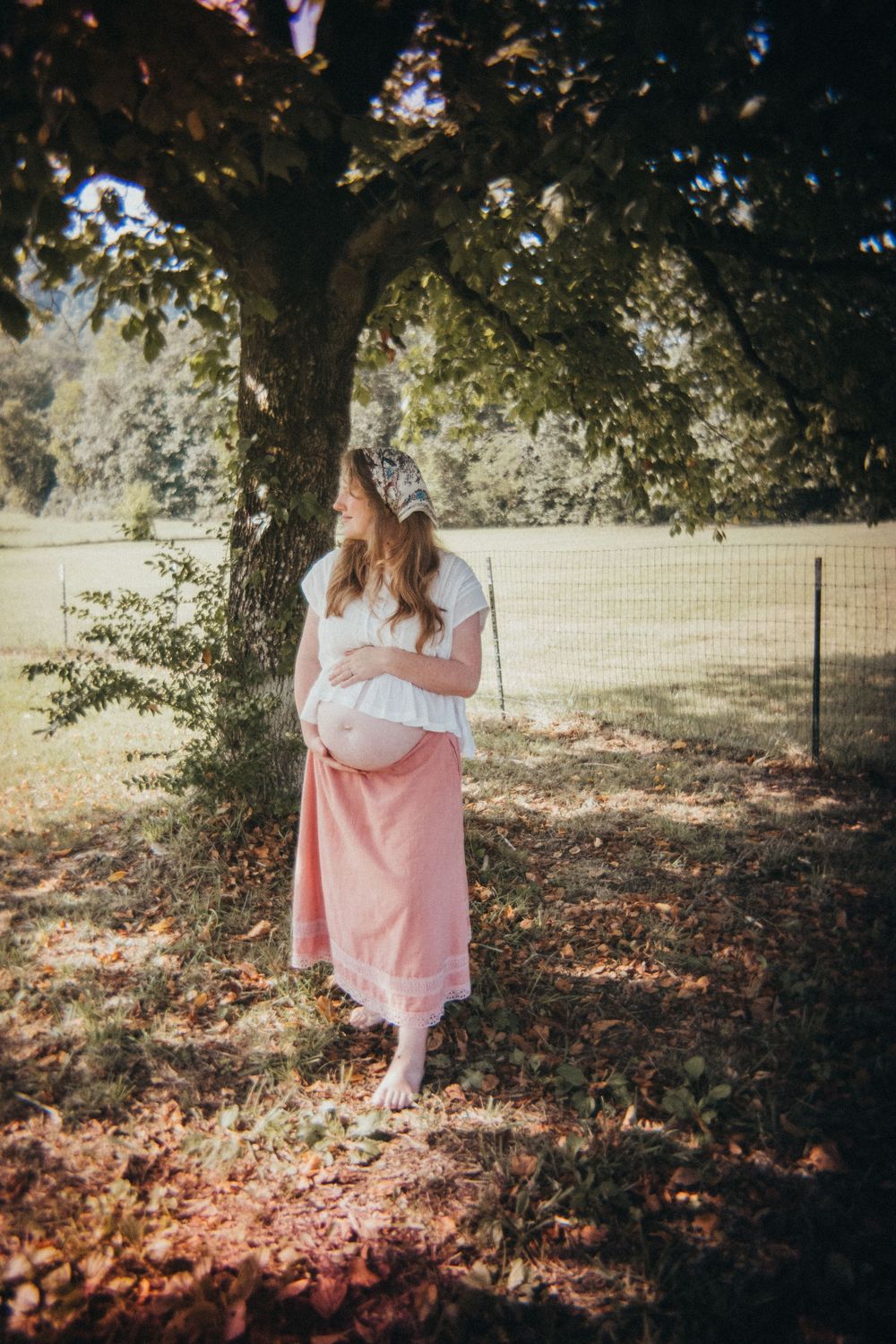 Pregnant woman in white top and pink skirt standing barefoot under a tree in a sunlit garden in Knoxville, TN.