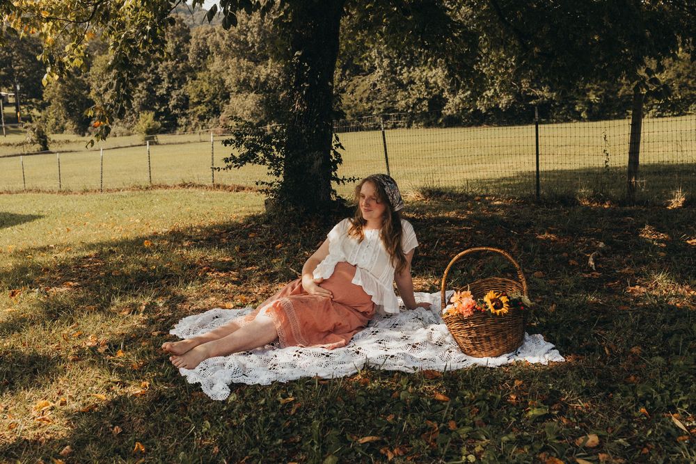 Woman in white top and pink skirt relaxing on lace blanket at park picnic with flower basket under tree in Knoxville, TN.
