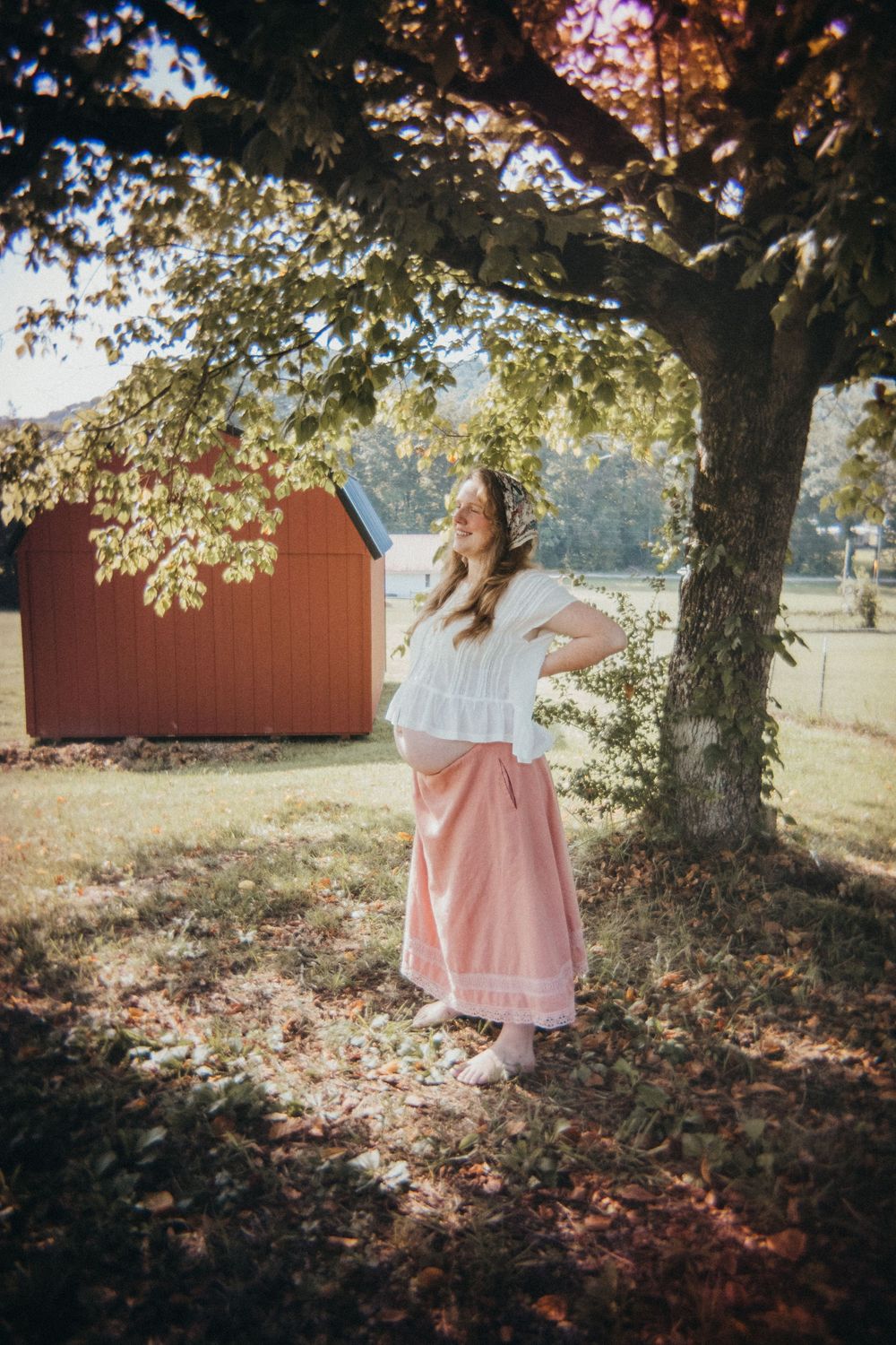Woman in white top and pink maxi skirt standing under a tree on a sunny farm with a red barn in the background in Knoxville, TN.