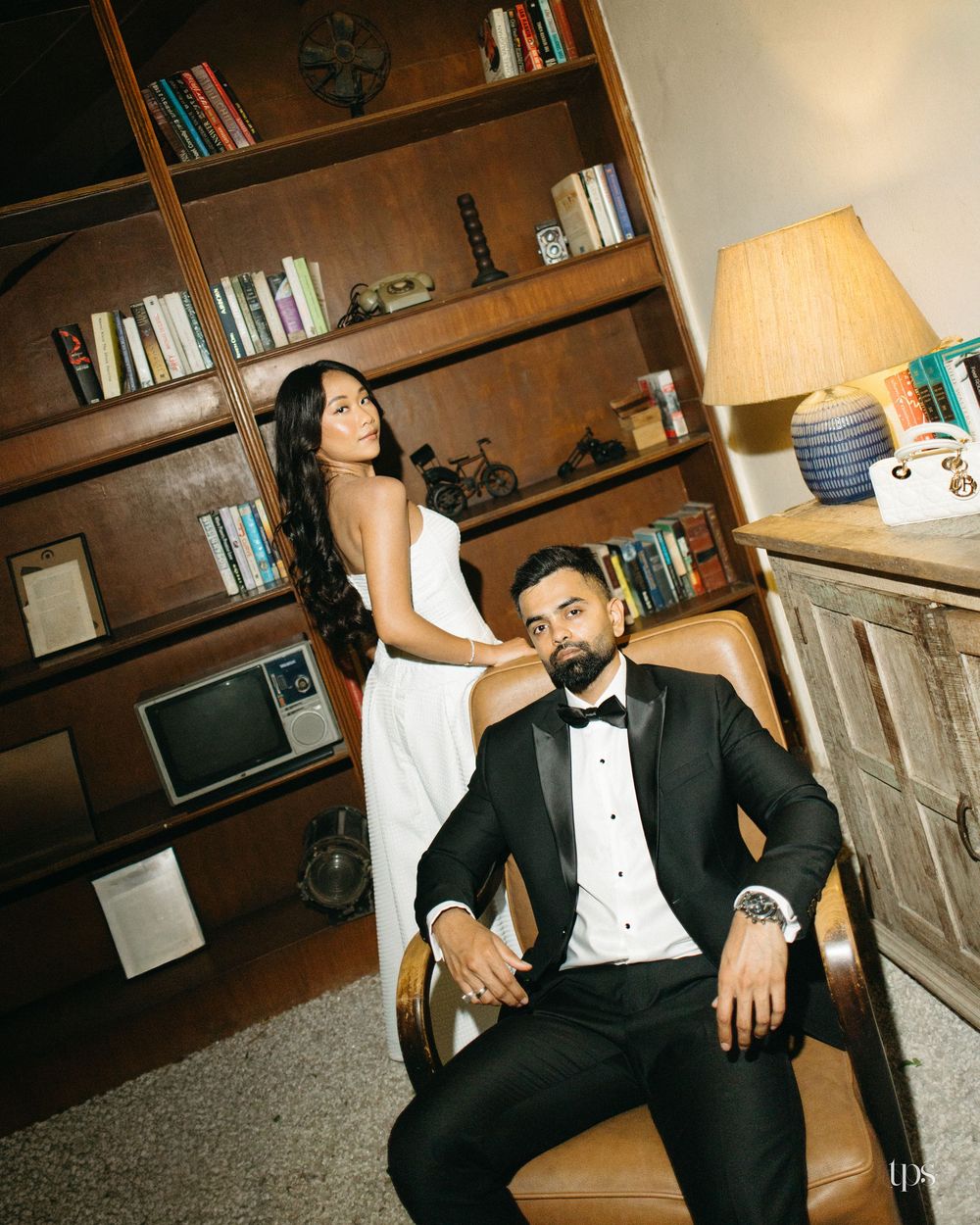 Elegant couple in formal attire posing in a library setting, man in tuxedo seated, woman in white gown standing behind.