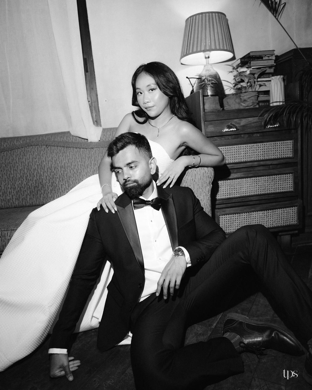 Black and white photo of a couple in formal wear, man in tuxedo seated on floor, woman in strapless gown posing behind him.