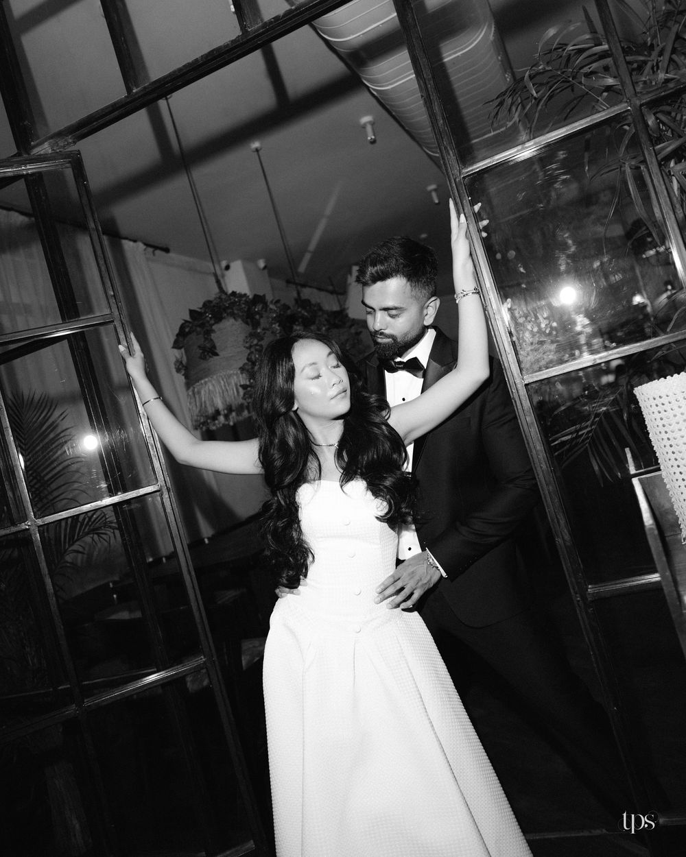 Black and white wedding photo of bride in white gown and groom in tuxedo posing elegantly by glass doors.