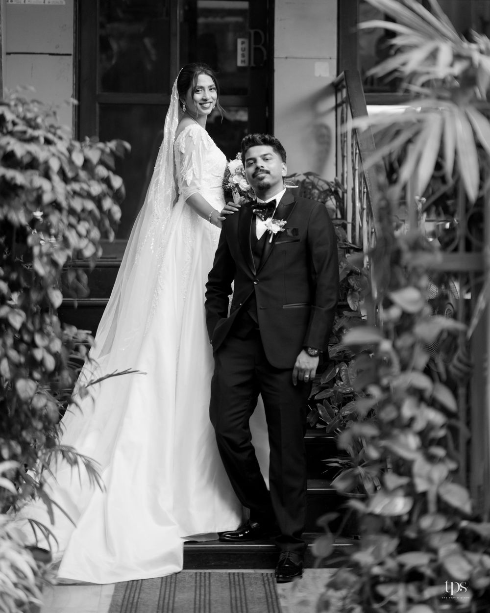 Black and white wedding photo of a bride in a veil and groom in a tuxedo posing among lush greenery.